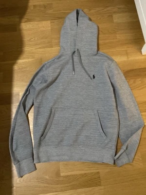 Grå hoodie från Ralph Lauren - Snygg grå hoodie från Ralph Lauren med klassisk huva och dragsko. Tröjan har känguruficka framtill och den ikoniska lilla Polo-loggan broderad på bröstet. Tillverkad i mjukt bomullsmaterial som är perfekt för chill dagar. Enkel och stilren design som funkar till det mesta.