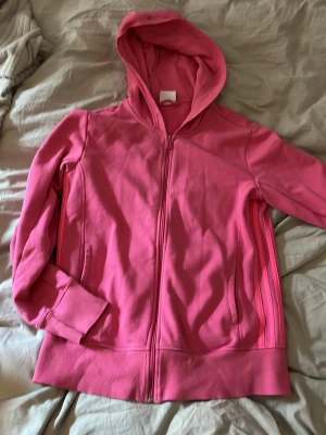 Rosa hoodie från Adidas, medium - Säljer en rosa hoodie från Adidas Sport Essentials i storlek medium. Hoodien har dragkedja framtill, två fickor och ribbade muddar. Tillverkad i mjukt material och har en klassisk huva.
