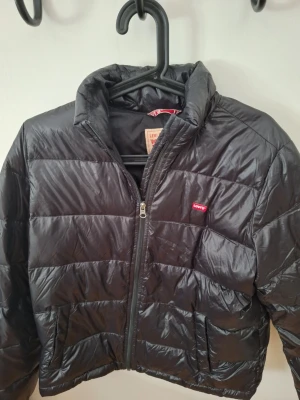 Svart dunjacka från Levi's - Snygg svart dunjacka från Levi's med quiltad design och dragkedja framtill. Jackan har en hög krage och två sidofickor. Klassisk Levi's-logga på bröstet och lätt glansigt material som ger en modern look.