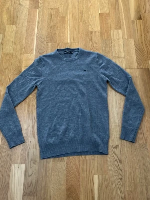 Grå rundhalsad merinoull tröja från J.Lindeberg - Snygg grå tröja från J.Lindeberg med rund halsringning och diskret logga på bröstet. Tröjan har långa ärmar och ribbade muddar vid ärmslut och nederkant. Perfekt för en stilren och grisch look. Tröjan är lite mindre i storleken, så passar nog xs också!