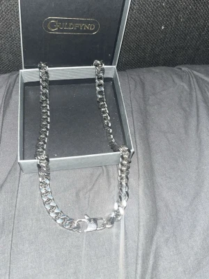 Kedjehalsband i silver från Guldfynd - Snyggt och stilrent halsband i silverfärgat metall från Guldfynd. Halsbandet har en bred, platt kedja med klassiskt karbinlås och kommer i originalask. Perfekt accessoar för att lyfta din outfit.