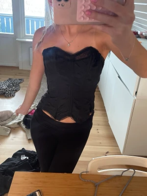 Svart spetsdetaljerad korsett - Säljer en svart korsett med spetsdetaljer upptill och formande sömmar. Modellen har en hjärtformad urringning och stängs med hyskor framtill. Materialet är glansigt och ger en snygg siluett. Perfekt för att lyfta vilken outfit som helst.