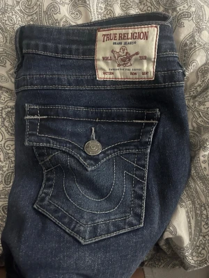 True Religion Joey Low Rise Flare Jeans - Säljer ett par mörkblå True Religion Joey jeans med låg midja och utsvängda ben. Klassisk kontrastsöm och stor ficka bak med ikonisk logga och knappdetalj. Tillverkade i slitstark denim, perfekta för dig som gillar en retro och avslappnad look.