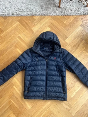 Polo Ralph lauren jacka  - Snygg mörkblå dunjacka från Polo Ralph Lauren med huva och dragkedja framtill. Jackan har två dragkedjeförsedda fickor och en liten röd logga på bröstet. Jackan är i storlek S och passar perfekt till slutet av vintern och våren som kommer! Ändå defekten på jackan ser man på bild 4 men det är inget man märker när man har på jackan. 