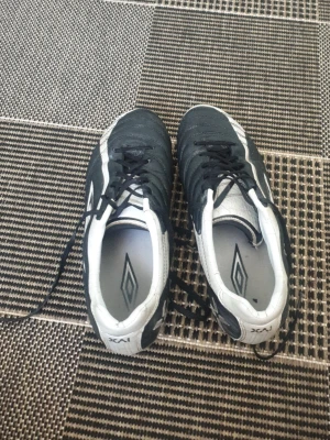 Svarta och vita Umbro fotbollsskor - Säljer ett par klassiska fotbollsskor från Umbro i svart och vitt. Skorna har snörning, låg profil och är tillverkade i syntetmaterial med Umbro-logga på sidan och innersulan. Perfekta för dig som vill ha stilrena och funktionella skor på planen.