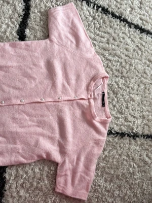 Ljusrosa kofta 💕 - Supersöt ljusrosa kofta från 157 med korta ärmar och pärlknappar framtill. Koftan är stickad i ett mjukt material och har ribbade kanter vid hals, ärmslut och nederkant. Perfekt för att addera en gullig touch till din outfit. OBS! Bild 2 och 3 saknar knappar på och första och sista hålen, hittar tyvärr inte reserv knapparna till! Använd 3 gånger men knapparna har råkat gått av så jag skulle säga att knapparna är väldigt sköra!