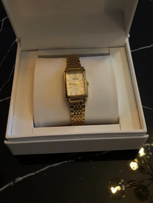 Trendig klocka  - Stilren guldfärgad klocka med fyrkantig urtavla och länkarmband i metall. Urtavlan är vit med tunna visare. Perfekt accessoar för att lyfta din outfit. Beteendet är slut och behövs bytas, vill man ha den endast som en assessor kan man skippa att byta batteri. Boxen följer ej med. Priset kan diskuteras! 