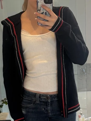 Svart kofta med röda och vita ränder - Svart stickad kofta med röda och vita ränder längs kanterna, ärmsluten och nederkanten. Koftan har en avslappnad passform och är öppen framtill utan knappar eller dragkedja. Perfekt att slänga över en topp för en snygg lager-på-lager-look.