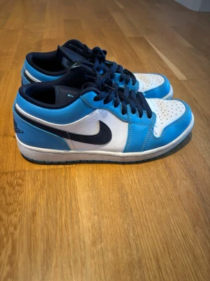 Air Jordan 1 Low blå och vita sneakers - Snygga Air Jordan 1 Low sneakers i blått och vitt med mörkblå detaljer och klassisk Nike Swoosh på sidan. Skorna har mörkblå snörning, vit tåbox med perforeringar och blå häl med Air Jordan-logga. Ovandelen är i läder och insidan är fodrad i mörkblå textil.