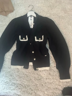 Svart kofta från Holly & Whyte - Stilren svart kofta från Holly & Whyte med vita kontrastdetaljer vid fickor, ärmslut och nederkant. Koftan har dekorativa knappar framtill och två fickor på bröstet med vita kanter och knappstängning. 