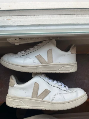 Vita Veja sneakers med beige detaljer - Säljer ett par klassiska vita Veja sneakers med beige mockadetaljer på sidorna och hälen. Tillverkade i läder och mocka för en stilren och clean look. Skriv om du har några frågor! 😊