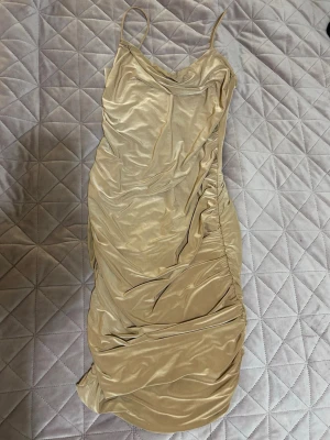 Champagnefärgad omlottklänning - Snygg champagnefärgad klänning med smala axelband och draperad omlott-design. Klänningen har en figurnära passform och är tillverkad i ett glansigt material som ger en lyxig känsla. Perfekt för dig som vill sticka ut med stil.