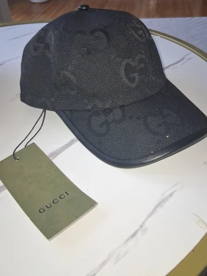Svart keps från Gucci med GG-mönster - Snygg svart keps från Gucci med klassiskt GG-monogram över hela kepsen. Kepsen har böjd skärm och justerbar passform. Tillverkad i textilmaterial med exklusiv känsla. Perfekt accessoar för att lyfta din streetwear-look. Har tags!