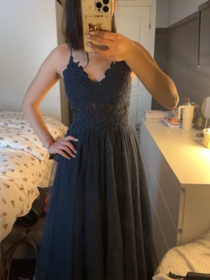 Marinblå spetsklänning med snörning - Elegant marinblå långklänning med spetsdetaljer och v-ringad hals. Klänningen har tunna axelband som korsas i ryggen och snörning baktill. Kjolen är i ett lätt, svepande material som ger en romantisk känsla. Perfekt för bal eller festliga tillfällen.