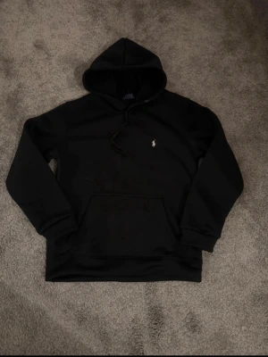 Svart hoodie från Polo Ralph Lauren - Klassisk svart hoodie från Polo Ralph Lauren med liten vit logga på bröstet. Hoodien har långa ärmar, huva med snörning och en stor magficka. Perfekt för en chill och stilren look.