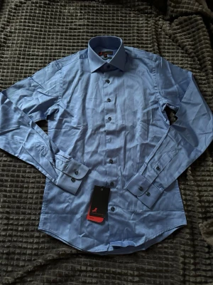 Ljusblå skjorta med knappar - Snygg ljusblå skjorta med klassisk krage och svarta knappar. Skjortan har lång ärm och är tillverkad i bomull, perfekt för en stilren look. Märke: Redmond.