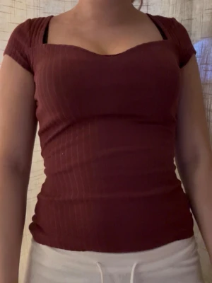 Vinröd ribbad topp med sweetheart-ringning - Snygg vinröd topp med ribbad struktur och korta ärmar. Toppen har en smickrande sweetheart-ringning och sitter tight mot kroppen. Perfekt att matcha med både jeans och kjol för en trendig look.