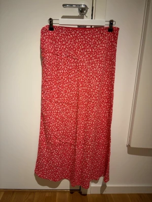 Röd blommig långkjol - Säljer en röd långkjol med små vita blommor. Kjolen har en rak och flowy passform som ger en avslappnad look. Perfekt för dig som gillar färg och mönster. Materialet känns lätt och skönt mot huden.