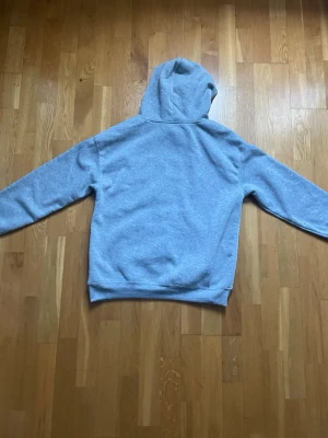 Ljusgrå hoodie med huva - En enkel och stilren ljusgrå hoodie med huva. Hoodien har långa ärmar och ribbade muddar vid ärmslut och nederkant. Perfekt för en avslappnad och bekväm look. Materialet ser ut att vara mjukt och skönt, troligen bomullsblandning.