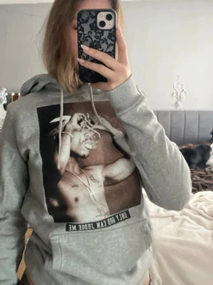 Grå hoodie med Tupac-tryck - Säljer en grå hoodie med stor svartvit bild av Tupac på framsidan och texten 'Only God Can Judge Me' under motivet. Hoodien har huva med dragsko och är tillverkad i mjukt sweatshirtmaterial. Perfekt för dig som gillar streetwear och hiphopstil.