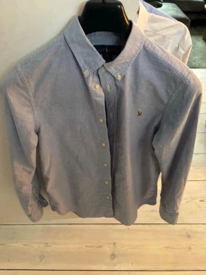 Ljusblå skjorta från Ralph Lauren - Klassisk ljusblå skjorta från Ralph Lauren med button-down krage och gul broderad logga på bröstet. Skjortan har långa ärmar, vita knappar och är tillverkad i bomull. Hör av dig vid frågor!