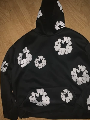 Svart hoodie med vita blommor - Säljer en svart hoodie från Denim Tears med stora vita blommönster tryckta över hela plagget, inklusive huvan och fickan framtill. Hoodien har en klassisk passform med huva och känguruficka. Perfekt för dig som vill sticka ut med en unik design.