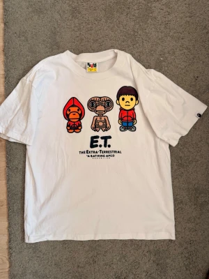 Vit E.T. t-shirt från BAPE - Vit t-shirt från A Bathing Ape med tryck av E.T. och två tecknade figurer på bröstet. Klassisk passform med korta ärmar och rund hals. Ikonisk BAPE-logga på ärmen. Perfekt för dig som gillar streetwear och filmklassiker.