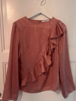 Rosa blus med volang från NA-KD - Supersnygg rosa blus från NA-KD i glansigt material. Blusen har långa ärmar och en dekorativ volang som går diagonalt över framsidan. Rund halsringning och knapp i nacken för enkel påklädning. Perfekt för dig som gillar trendiga detaljer.
