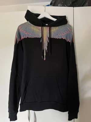 Svart hoodie med färgglada vingar - Säljer en svart hoodie från Marcelo Burlon med ikoniskt färgglatt vingmönster över axlar och bröst. Hoodien har huva med dragsko och en stor magficka. Perfekt för dig som vill sticka ut med en unik och trendig design.