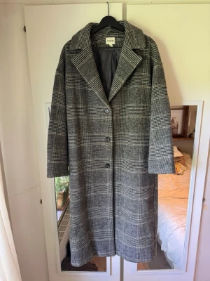Grå rutig kappa från Kappahl  - Stilren grå kappa från Kappahl med klassiskt rutigt mönster i svart, grått och brunt. Oversized passform, bred krage och tre knappar framtill. Fodrad insida för extra komfort. Perfekt för dig som gillar en trendig och tidlös look.