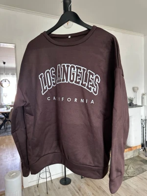 Brun sweatshirt med Los Angeles-tryck - Cool brun sweatshirt med texten 'Los Angeles California' i vitt framtill. Tröjan har rund halsringning och långa ärmar. Perfekt för en avslappnad streetwear-look och passar till både jeans och mjukisbyxor.
