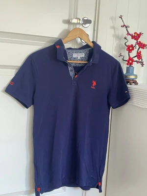 Marinblå pikétröja från U.S. Polo Assn. - Marinblå pikétopp från U.S. Polo Assn. med korta ärmar, klassisk krage och orange broderad logga på bröstet. Insidan av kragen har ett blommigt mönster och toppen är tillverkad i mjuk bomull. Perfekt för en sportig och stilren look.