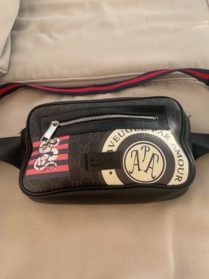 gg axelväska med tryck och logga - Snygg axelväska från gg i svart med GG-monogram, färgglada detaljer och texttryck. Väskan har en justerbar axelrem i rött och blått, en framficka med dragkedja och coola grafiska prints. Tillverkad i canvas och läder för en exklusiv känsla.