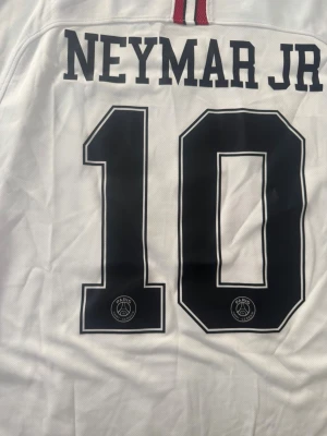 PSG Neymar Jr matchtröja vit - Säljer en vit PSG matchtröja med Neymar Jr och nummer 10 på ryggen. Tröjan har Champions League-märke på ärmen och är tillverkad i ett lätt, ventilerande material. Perfekt för dig som älskar fotboll och vill bära din favoritspelare på ryggen.