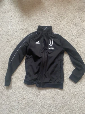 Svart Juventus träningsjacka Adidas - Snygg svart träningsjacka från Adidas med Juventus och Jeep-logga på bröstet. Jackan har hel dragkedja, hög krage och vita detaljer längs ärmarna. Perfekt för fotbollsträning eller chill. Tillverkad i polyester.