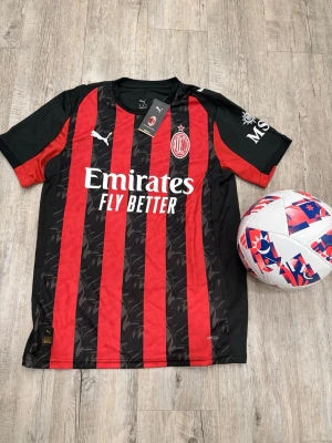 AC Milan matchtröja Puma röd/svart - AC Milan matchtröja från Puma i rött och svart med klassiska vertikala ränder och klubbmärke på bröstet. Tröjan har korta ärmar, rund hals och tryckt sponsorlogga framtill. Tillverkad i lätt och ventilerande material, perfekt för fotbollsträning.