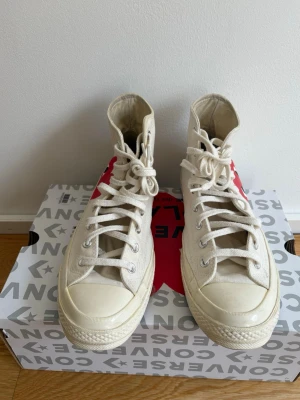 Converse x CDG Play vita high tops - Snygga vita Converse x Comme des Garçons Play high tops med det ikoniska röda hjärtat på sidan. Klassisk canvas, vita snören och gummisula. Perfekta för dig som vill ha en stilren sneaker med en twist från ett populärt samarbete.