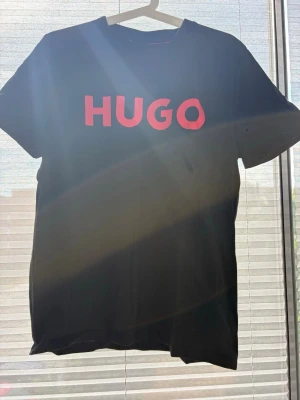 Svart HUGO t-shirt med röd logga - Snygg svart t-shirt från HUGO med stor röd logga framtill. Klassisk rund halsringning och korta ärmar. Tillverkad i mjuk bomull som känns skön mot huden. Perfekt för en clean och stilren look.