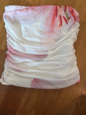 Vit bandeau tubtopp med blommigt mönster - Säljer en vit bandeau tubtopp med blommigt mönster i rosa och röda toner. Toppen är ärmlös och har rynkade sidor för en snygg passform. Perfekt för dig som vill ha en fräsch och somrig look.