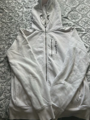 Vit hoodie från Sail Racing - Säljer en vit hoodie från Sail Racing med dragkedja framtill och broderad logga på bröstet. Hoodien har en extra dragkedjeficka på bröstet och justerbar huva med snören. Perfekt för dig som gillar stilren och sportig design.