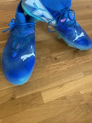 Blåa Puma fotbollsskor med dobbar - Säljer ett par blåa fotbollsskor från Puma med vita detaljer och logga. Skorna har snörning, stickad strumpkrage och dobbar i ljusgrönt. Ovandelen är i syntetmaterial med små vita prickar för extra grepp på planen. Perfekta för dig som vill sticka ut på fotbollsplanen. Storlek 40,5 ,använda en säsong jätte bra skicka bara lite smutsiga😁😁😁orginal pris 1170kr😁