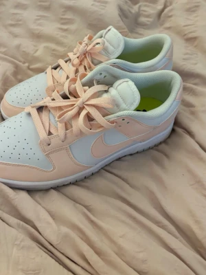 Nike Dunk Low i vitt och ljusrosa - Nike Dunk Low sneakers i vitt och ljusrosa läder med klassisk swoosh på sidan. Skorna har perforerad tå, vita sulor och matchande ljusrosa skosnören. Insidan är vadderad för extra komfort och designen är stilren och trendig.