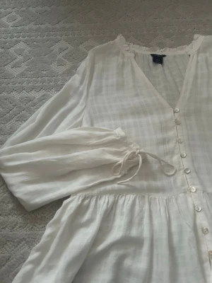 Vit blus med knappar och volang - Söt vit blus från H&M med v-ringning, knappar framtill och volang nedtill. Blusen har långa ärmar med knytband vid ärmsluten och är tillverkad i ett lätt, luftigt tyg med diskret rutmönster. Perfekt för en trendig och avslappnad look.