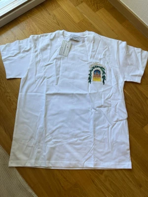 Vit Casablanca t-shirt med tryck - Säljer en vit t-shirt från Casablanca med färgglatt tryck av en byggnad och texten 'Avenida Casablanca' på ryggen. T-shirten har korta ärmar och är gjord i mjuk bomull. Perfekt för dig som gillar unika prints och streetwear vibes.