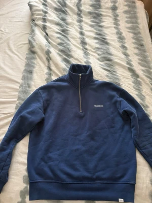 Blå half zip tröja från Les Deux - Snygg blå sweatshirt med half zip och hög krage från Les Deux. Tröjan har broderad logga på bröstet och ribbade muddar vid ärmslut och nederkant. Perfekt för en avslappnad och stilren look.