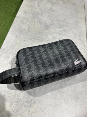 Svart necessär från Lacoste - Snygg svart necessär från Lacoste med geometriskt mönster och klassisk krokodil-logga. Väskan har dragkedja, ett praktiskt handtag på sidan och en extra ficka på baksidan. Tillverkad i tåligt konstläder som ger en stilren look.
