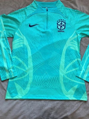 Nike Brasil långärmad träningströja - Snygg långärmad träningströja från Nike med Brasil-logga. Tröjan är turkos med ljusgröna grafiska mönster och har halv dragkedja framtill. Perfekt för fotbollsträning eller sportiga aktiviteter. Materialet är lätt och andas bra.