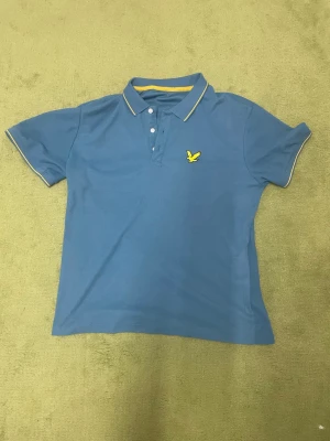 Blå pikétröja från Lyle & Scott - Snygg blå pikétröja från Lyle & Scott med klassisk krage, tre knappar och gul logga på bröstet. Tunn gul kant på krage och ärmslut ger extra stil. Perfekt till jeans eller shorts.