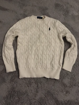 Vit kabelstickad tröja Polo Ralph Lauren - Kabelstickad vit tröja från Polo Ralph Lauren med klassisk rund halsringning och svart broderad logga på bröstet. Tröjan har ribbade muddar vid ärmslut och nederkant, perfekt för en stilren och tidlös look.