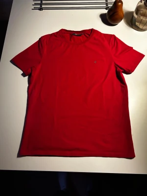 Röd t-shirt med broderad detalj - Snygg röd t-shirt i bomull med korta ärmar och en liten broderad lilja på bröstet. Enkel och stilren modell som passar till det mesta. Perfekt för dig som gillar klassiska plagg med en twist.